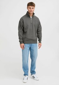 Sudadera gris con cremallera y cuello alzado, con puños y dobladillo de canalé, combinada con jeans azul claro y zapatillas blancas. Presenta un logo bordado.