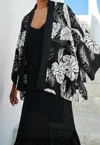 Lekki kimono w kolorze czarnym z dużymi białymi wzorami liścia monstera. Posiada szerokie rękawy i jednolity czarny wyko�ńczenie, noszony na czarnej sukience.