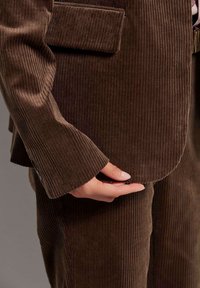 Braunes Cordanzug mit vertikaler Rippstruktur; verfügt über ein tailliertes Jacket und weit ausgestellte Ärmel, eine Hand hält den Saum des Jackets.