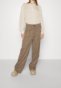 Žena stojící v krémové plisé haleně s knoflíky, hnědých kostkovaných širokých kalhotách a béžových objemných botách na bílém pozadí.