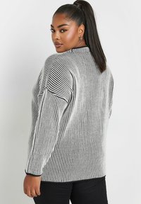 Maglione a righe bianche e nere a maglia con maniche lunghe, colletto alto e texture a coste, abbinato a pantaloni neri.