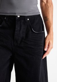 Black denim pants featuring a relaxed fit, subtle frayed edges on pockets, and a silver button closure. Texture appears soft and worn.