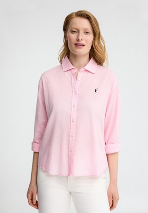 Kvinde med rødt hår iført en lyserød skjorte med knapper og opkørte ærmer samt hvide bukser, stående foran en hvid baggrund.