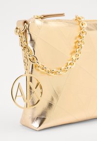 Złota metaliczna torebka kopertowa z geometrycznym wzorem, z łańcuszkowym paskiem i okrąłym charms z logo. Gładka tekstura z błyszczącym wykończeniem.