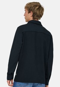 Junger Mann mit kurzen blonden Haaren, der ein schwarzes, langärmeliges Hemd mit Kragen und blaue Jeans trägt, von hinten vor einem einfarbigen Hintergrund.