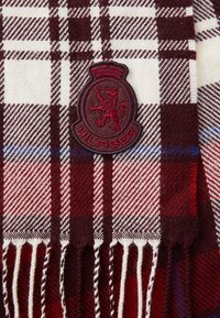 Czerwono-burgundowa kraciasta szalik wykonany z miękkiego materiału z białym frędzlem; posiada haftowaną w kolorze bordowym łatkę "Hilfiger" z emblematem lwa.