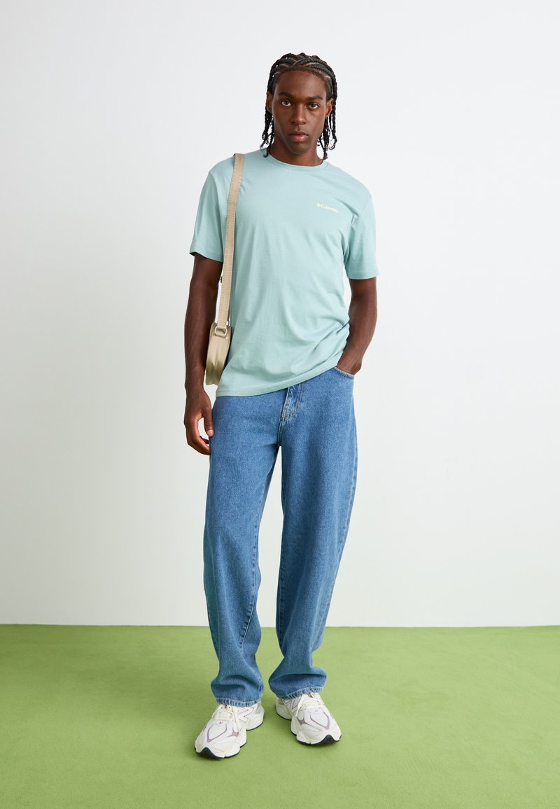 T-shirt en coton bleu clair avec un col rond, associé à un jean en denim ample. Le mannequin porte des baskets blanches et violettes ainsi qu'un sac à épaule beige.