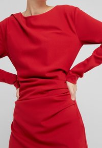 Rotes langärmliges Kleid aus glattem Stoff, mit Rundhalsausschnitt, geraffter Taille und figurbetonender Silhouette mit sanften Drapierungen.