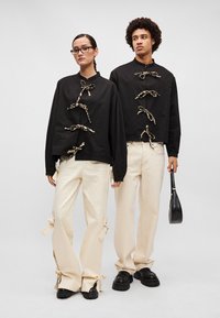 Juodas trumpas sijonas su auksiniais akcentais ir raišteliais, derinamas su kreminės spalvos plačiomis kelnėmis su kaspinais prie siūlių. Abu vilki juodus batus.