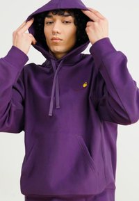 Lilla hoodie lavet af blødt stof med en frontlomme, justerbar snoretræk og et lille gult logo på brystet.