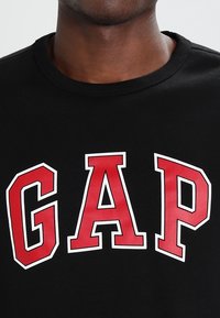 Camiseta de manga corta negra con letras rojas "GAP" en negrita y bordes blancos. Textura de algodón suave, diseño de cuello redondo.