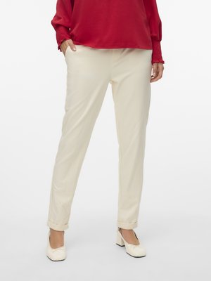 Ženska v kremnih hlačah z ravnimi hlačnicami in zavihanimi robovi, obuta v krem čevlje z blok peto ter v rdečo bluzo z dolgimi rokavi, z rokami v žepih.