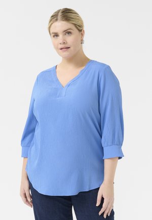 Blusa de color azul cielo, texturizada, con escote en V y mangas de tres cuartos. Presenta un corte relajado y un sutil detalle de costura a lo largo de los bordes.