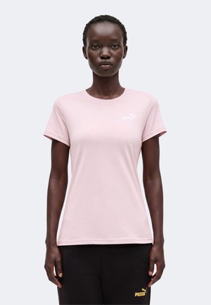 Γυναίκα με ανοιχτό ροζ μπλουζάκι Puma και μαύρο παντελόνι Puma στέκεται μπροστά σε απλό φωτεινό φόντο.