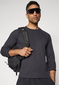 Schwarzes strukturiertes Langarmshirt mit Rundhalsausschnitt, getragen mit einem schwarzen Rucksack mit verstellbaren Trägern. Das Modell trägt eine dunkle Sonnenbrille.