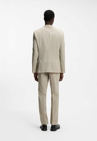 Hombre de pie de espaldas, vistiendo un traje beige con zapatos negros, sobre un fondo blanco liso.