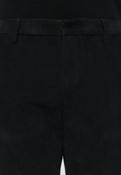 Pantalones de pana negros con una superficie texturizada, corte recto y lazos para el cinturón estándar. Vista en primer plano de la cintura y la tela.