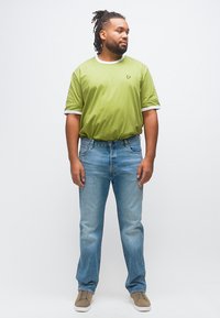 Άνδρας με ντρέντλοκς που φοράει ανοιχτό πράσινο T-shirt με λογότυπο πεταλούδας, μπλε τζιν και μπεζ αθλητικά παπούτσια, στέκεται με τα χέρια χαλαρά.