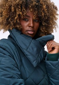 Gequilted teal jas met een hoge kraag, voorzien van diamantpatronen en contrasterende groene manchetten. Gladde textuur en puffy ontwerp.