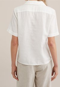 Vit linneskjorta med korta ärmar och krage, med en avslappnad passform och en något texturerad yta. Bärs med ljusa byxor.