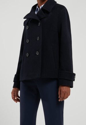 Person trägt einen marineblauen doppelt geknöpften Pea Coat mit geknöpften Manschetten über einem hellblauen Hemd mit Kragen und marineblauen Hosen.