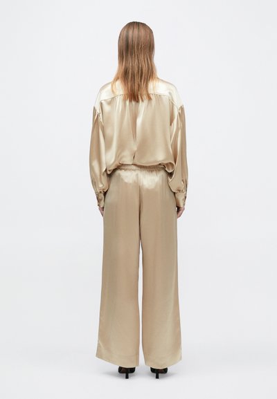 Bézs szatén blúz és bőszárú nadrág, mindkettő sima textúrájú és laza szabású. A nadrág rugalmas derékpánttal rendelkezik; a felső leengedett vállakkal van kialakítva.