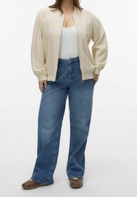 Beige bomberjacka med lös passform, rund krage och samlade ärmar. Matchad med högmidjade blåjeans och bruna skor.