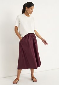 Witte T-shirt met korte mouwen gecombineerd met een knielange maroonrok met een elastische taille en zijzakken, aangevuld met bruine sandalen.