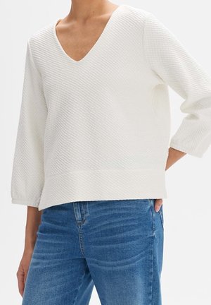 Kvinde iført tekstureret hvid V-hals sweater med trekvart ærmer og blå denim jeans, hånd i lommen, mod hvid baggrund.