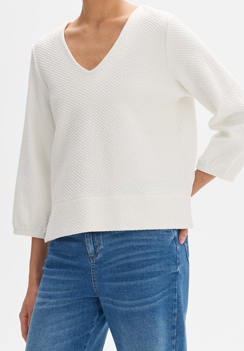 Femme portant un pull blanc texturé à col en V avec manches trois quarts et jean en denim bleu, main dans la poche, sur fond blanc.