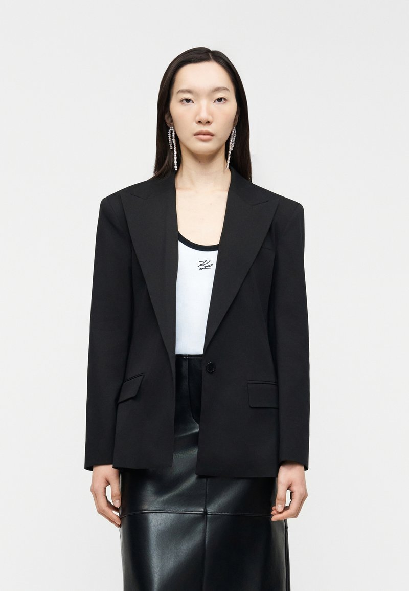 Γυναίκα με μακριά σκούρα μαλλιά φοράει υπερμεγέθη μαύρο σακάκι, λευκό τοπ με λογότυπο, μαύρη δερμάτινη φούστα και μεγάλα κρεμαστά σκουλαρίκια, κοιτάζοντας μπροστά.