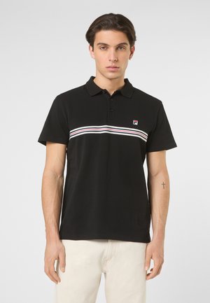 Uomo in piedi che indossa una polo nera con strisce orizzontali bianche, rosse e blu scuro e pantaloni chiari, su uno sfondo semplice.