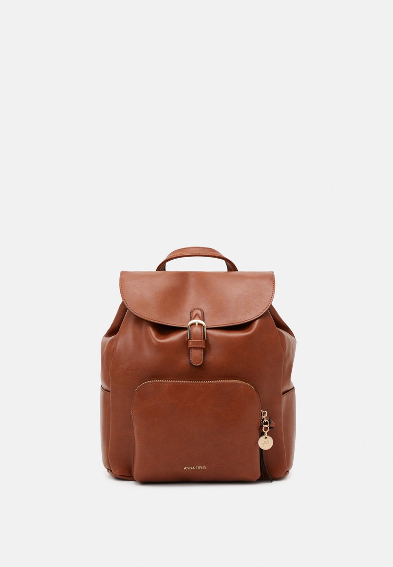 Sac à dos en cuir marron avec poche avant zippée, rabat supérieur sécurisé par boucle et petite poignée sur un fond clair uni.