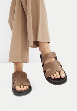 Brązowe skórzane klapki na płaskiej podeszwie z dwoma szerokimi paskami z wycięciami, teksturowaną czarną podeszwą i casualowym designem odpowiednim na ciepłą pogodę.