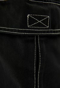 Svart denimtyg med kontrasterande vit sömnad. Designen har en rektangulär etikett med ett broderat korsmönster.