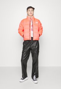 Giubbotto imbottito arancione con colletto alto, chiusura con zip e dettagli neri, abbinato a pantaloni in ecopelle neri e sneaker bianche.
