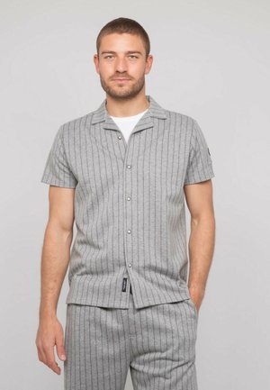 Homme aux cheveux courts et barbe portant une chemise à manches courtes grise à fines rayures boutonnée et un pantalon assorti sur un fond uni.