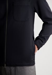 Chaqueta navy de cremallera con tejido texturizado, que presenta un bolsillo en el pecho y bolsillos laterales. Incluye un pequeño logotipo cerca del dobladillo.