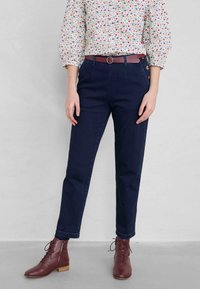 Navyblauwe denim broek met een rechte pasvorm, voorzien van bronzen knopen en een riem met een ronde gesp. Gecombineerd met een bloemenblouse en bruine veterschoenen.
