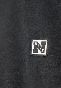 Melnas audums ar gludu faktūru, kurā ir balta Nike logo uzlīme apakšējā labajā stūrī, kvadrāta formā ar kontrastējošiem melniem elementiem.