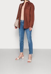 Camicia marrone a bottoni sopra una maglietta rosa chiaro, jeans skinny blu e sandali alti intrecciati beige. Borsa semplice in beige.