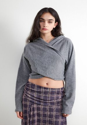 Junge Frau mit langen dunklen Haaren, die einen grauen kurzen Wickelpullover und einen violetten karierten gerafften Rock trägt, steht vor einem weißen Hintergrund.