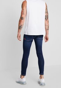 Jeans ajustados de mezclilla en azul oscuro con una textura suave, que presentan un discreto parche de logo rojo en el bolsillo trasero. Combinados con una camiseta sin mangas blanca.