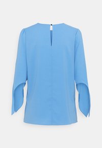 Blouse bleu clair à manches longues évasées avec une petite ouverture en forme de goutte dans le dos, fermée par un bouton noir.