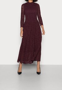 Burgundy spets maxiklänning med långa ärmar, hög halsringning och intrikata blommönster; kombinerad med svarta spetsiga klackar.