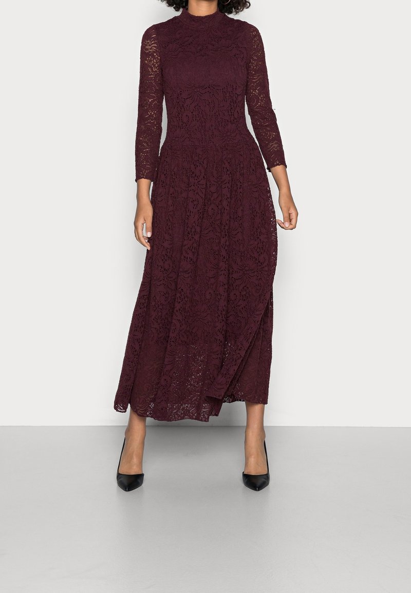 Burgundy spets maxiklänning med långa ärmar, hög halsringning och intrikata blommönster; kombinerad med svarta spetsiga klackar.