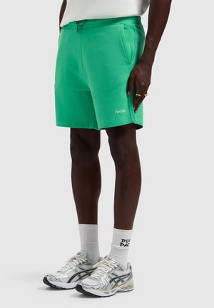 Man met felgroene Pure Path shorts, witte sokken met tekst en zilveren hardloopschoenen, staand tegen een effen achtergrond.