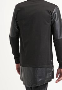 Top nero a maniche lunghe con corpo in cotone e maniche effetto pelle. Presenta un orlo curvo e dettagli con zip laterale. Tessuto vellutato.