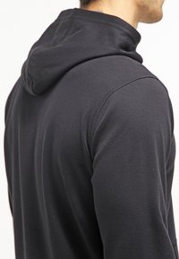 Homme portant un sweat à capuche noir vu de dos et de côté, avec un focus sur les détails des épaules et de la capuche.