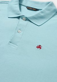 Lichtblauwe polo gemaakt van textuurstof, met een drie-knopen sluiting en een klein rood geborduurd logo op de borst.